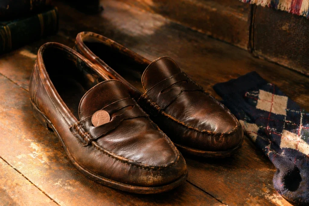 worn brown penny loafers with coin on wooden floor beside argyle socks
